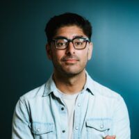 A headshot of Kashif Shaikh, co-founder of Pillars Fund. He wears a casual blue button-down shirt and dark-rimmed glasses. He is photographed in front of a neutral, dark-colored background.
