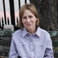 Portrait of director Kelly Reichardt. The image depicts a blonde middle aged woman sitting on a bench in front of a tree and a metal fence. She is smiling and wears a purple fleece button-up shirt.