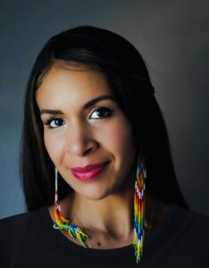 A photo of Kiley Acosta from the Spanish and Portuguese department at UCSB. Acosta appears against a neutral blue background, wearing a black shirt and long beaded earrings. She has long black hair and is smiling.