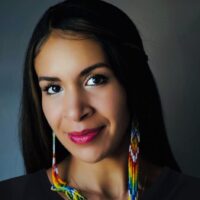 A photo of Kiley Acosta from the Spanish and Portuguese department at UCSB. Acosta appears against a neutral blue background, wearing a black shirt and long beaded earrings. She has long black hair and is smiling.