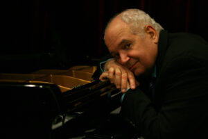 MichaelMortilla_Headshot A headshot of pianist Michael Mortilla. The image depicts a man leaning over an open grand piano with his head on his hands. He is smiling and looking thoughtfully towards the camera. He wears a black blazer and a blue button up shirt