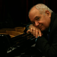 A headshot of pianist Michael Mortilla. The image depicts a man leaning over an open grand piano with his head on his hands. He is smiling and looking thoughtfully towards the camera. He wears a black blazer and a blue button up shirt