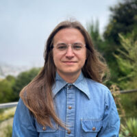 Alex Lilburn, a graduate student in Film and Media Studies at UCSB, stands against a background of green trees. He has glasses and long brown hair, and is wearing a blue buttoned shirt.