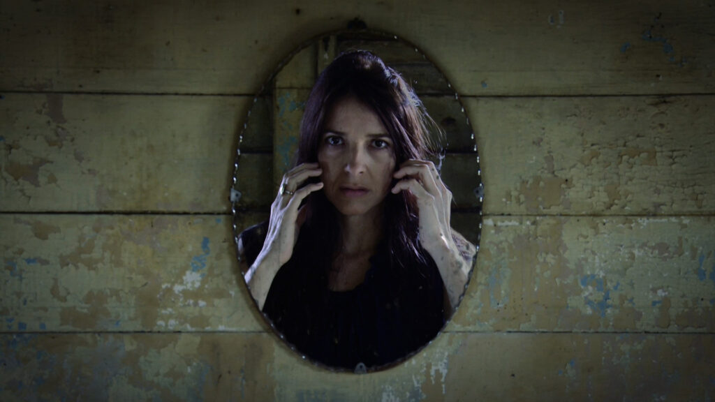 A woman looks in a mirror that is hung on a wall made of horizontal weathered wooden boards. She holds her hands to her cheeks and looks upset.
