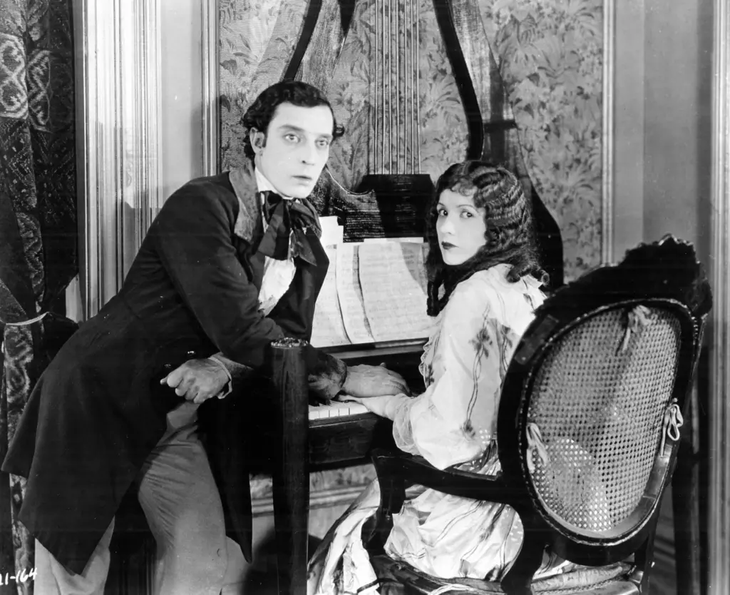 This image is a black and white promotional still from the Buster Keaton film Our Hospitality. The image depicts actor/director Buster Keaton leaning on a piano with a hand placed over actor, Natalie Talmadge's hand as she sits in front of the piano on a chair. They are dressed in upper middle class outfits from the 1920s. The piano has many pages of sheet music propped up on the stand. They both are not smiling, facing towards the camera.