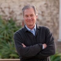 Headshot of screenwriter, Peter Seaman. He wears a sweater overtop a blue button up shirt. He is posed with his arms cross, smiling towards the camera, against the exterior of a building with plants in front of it.