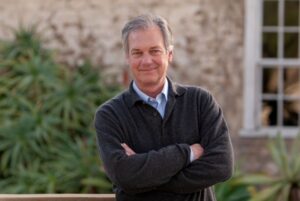 PeterSeaman_Headshot_2 Headshot of screenwriter, Peter Seaman. He wears a sweater overtop a blue button up shirt. He is posed with his arms cross, smiling towards the camera, against the exterior of a building with plants in front of it.