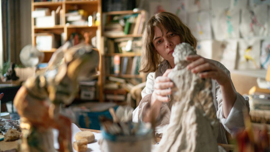 Still from the film, Showing Up. The image depicts a woman with short hair in an art studio. She has her hands on a clay sculpture and is molding details. The background of her studio is blurred and sees various shelves with books and other materials. The walls are lined with drawings and in the foreground there is a finished sculpture.