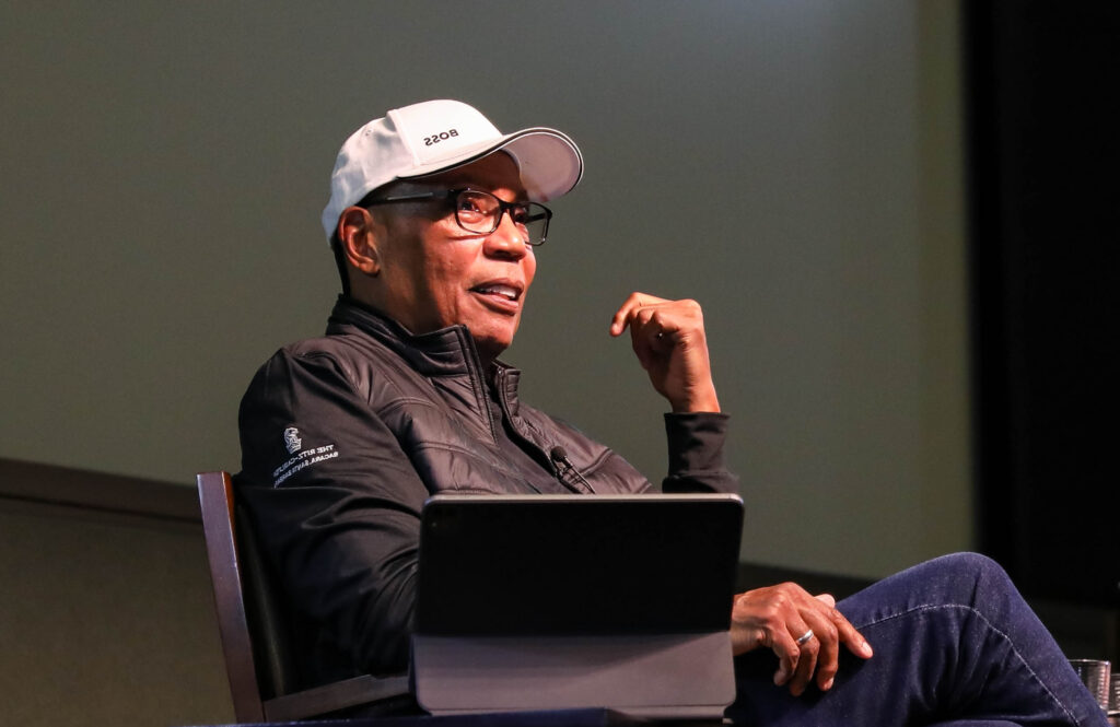 Director Paris Barclay addresses the Pollock Theater audience from the stage. He wears a white baseball cap, glasses, and a black jacket. He is smiling and gesturing with his left hand.
