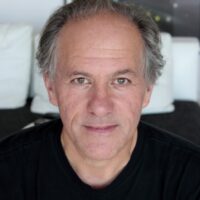 George Legrady, professor of Media Arts & Technology at UCSB, poses against a light background. He has grey hair and is smiling. He wears a black shirt.