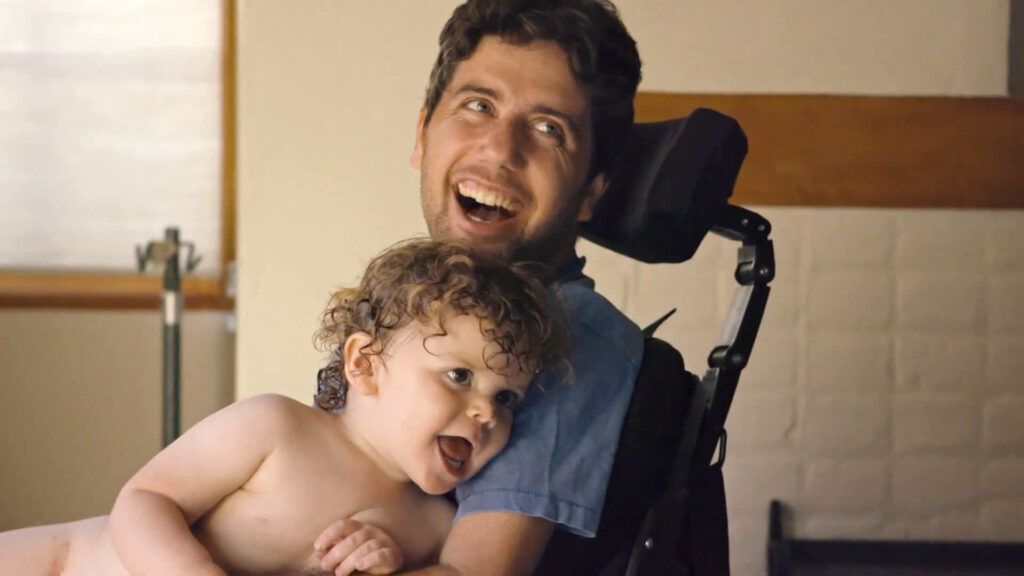 This image depicts a man smiling wearing a blue shirt smiling and holding an infant. He sits in a wheelchair and looks away from the camera, appearing to be laughing.