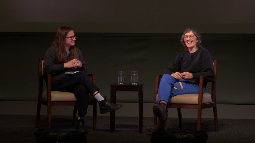 Stage photo of Moderator Alex Lilburn and Deborah Stratman on stage at the UCSB Pollock Theater. Deborah(right) appears smiling in a grey sweater, blue pants, brown rimmed glasses and is smiling towards the audience. Alex (left) has long hair, glasses, black slacks and a grey button up shirt, he is smiling and facing Deborah.