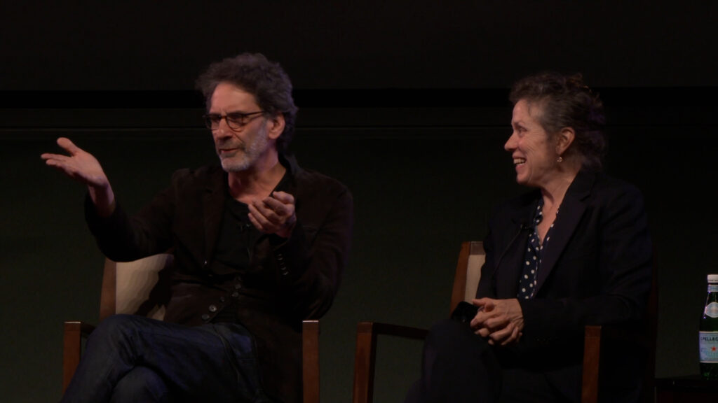 Guests Joel Coen and Frances McDormand on stage during a Q&A at the Pollock Theater. Joel, on the left, gestures with his hands, while Frances, on the right, smiles at him.