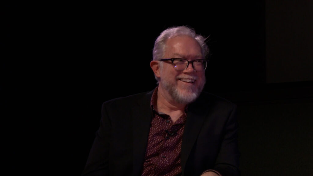 Moderator Jim Kearney smiling during a Q&A at the Pollock Theater. He is wearing a black blazer over a maroon polka dot dress shirt.