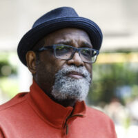 Filmmaker Sam Pollard appears against a light background looking toward the right side of the frame. He is wearing a black hat, black glasses, and an orange jacket.