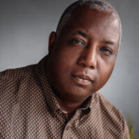 A headshot of director and producer Kemp Powers. He poses against a neutral background and wears a black and brown patterned button up shirt.