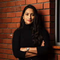 Director/producer Sreemoyee Singh stands against a brick wall. She is wearing a black turtleneck and smiling at the camera.