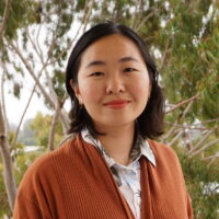 English professor Heidi Amin-Hong stands against a background of green leafy trees. She is smiling, and wearing an orange cardigan sweater.