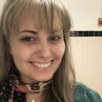 PhD student Kyna McClenaghan stands against a white tile background. She is smiling at the camera, and wearing a green sweater and a printed scarf.