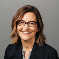 Filmmaker Nicole Newnham appears against a neutral background. She is smiling, and wears glasses, a black blazer with a print blouse.