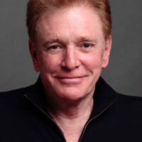 Actor William Atherton appears against a neutral grey background. He is smiling and wearing a black open-collared shirt.