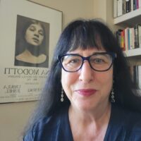 Patrice Petro, the Dick Wolf Director of the Carsey-Wolf Center appears against a background of a bookshelf and a poster featuring Tina Modotti. She is wearing a black v-neck blouse, glasses, and dangly earrings.