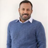 Actor Rizwan Manji stands against a neutral beige background. He is smiling and is wearing a blue sweater and a white collared shirt.