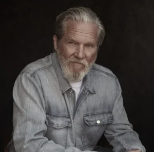 Jeff Bridges_HeadShot Headshot of actor Jeff Bridges. He is pictured in front of a textured black background, and is wearing a light wash denim jacket and a white t-shirt.