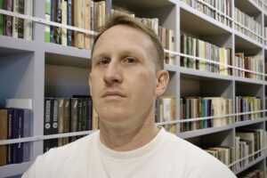 ClintTerrell_headshot Head shot of moderator and PHD candidate Clint Terrell. He is pictured wearing a white t-shirt and is standing in front of a shelf of books.