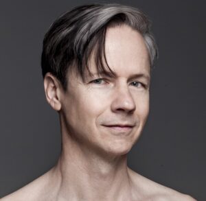 Headshot of filmmaker John Cameron Mitchell. He is pictured from the shoulders up and shirtless and is smiling slightly in front of a grey back drop.