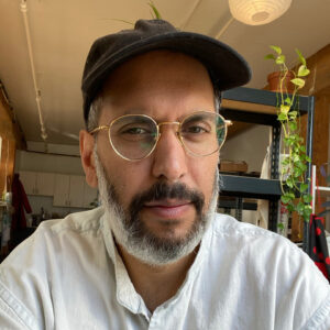 Head shot selfie of Sameer Farooq. He is pictured in a black cap, and round gold rimmed glasses in front of what appears to be a kitchen.