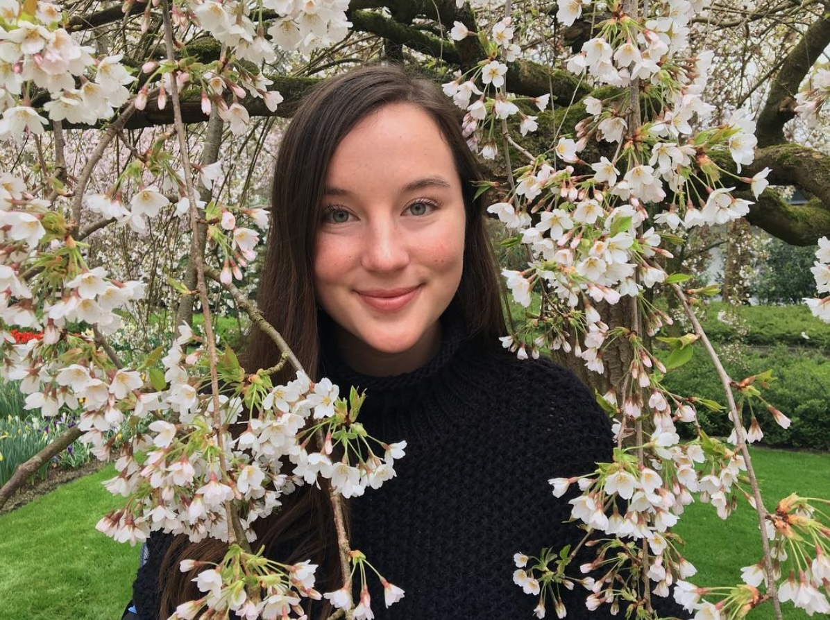 Headshot of UCSB Alum Elizabeth Cook. She is pictured smiling and standing between the branches of a flowering tree, in front of a mossy garden and lake.