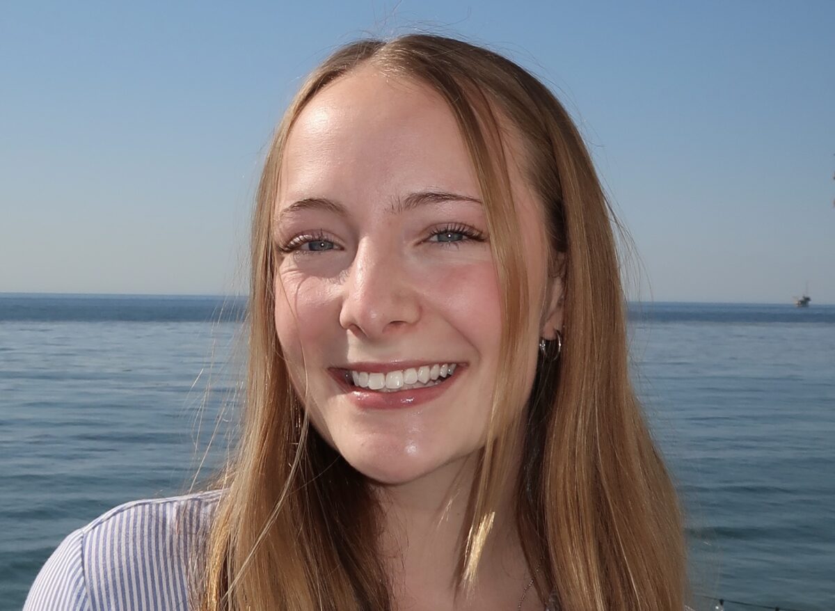 Close up headshot of student, Isabel Santiago. She has warm-toned blonde hair and blue eyes. She is smiling and wearing a purple top, she is standing in front of the ocean.