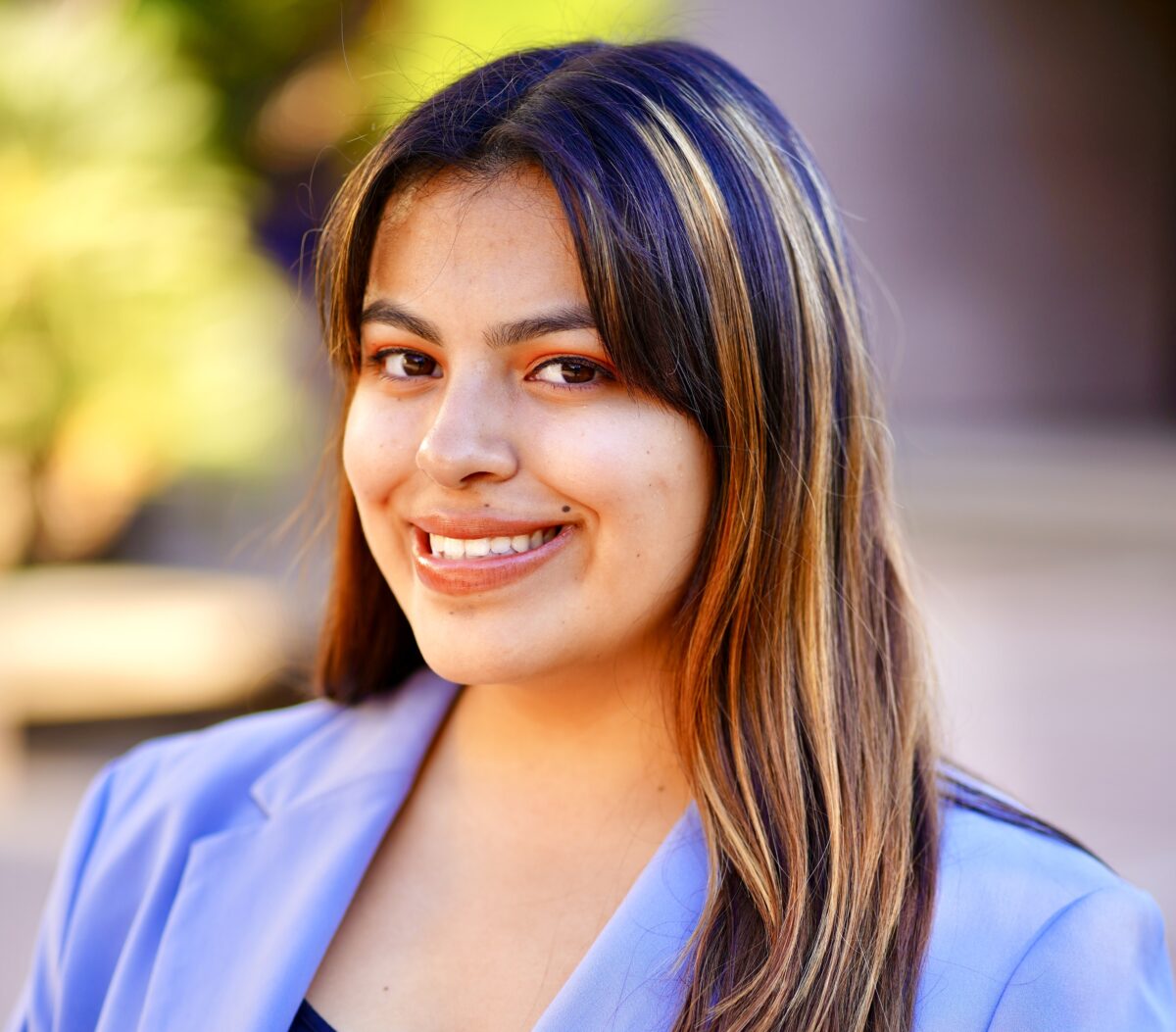 A headshot of Liliana Linan, a student of communication and Film and Media Studies. She is smiling at the camera. She has brown hair with blonde streaks and is wearing a sky blue blazer.