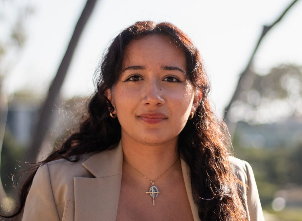 Headshot of student journalist Michelle Cisneros. She is pictured in front of a blurry nature background. She is wearing a beige blazer, gold earrings, a nose piercing, and a necklace with a gold cross and silver pendant.
