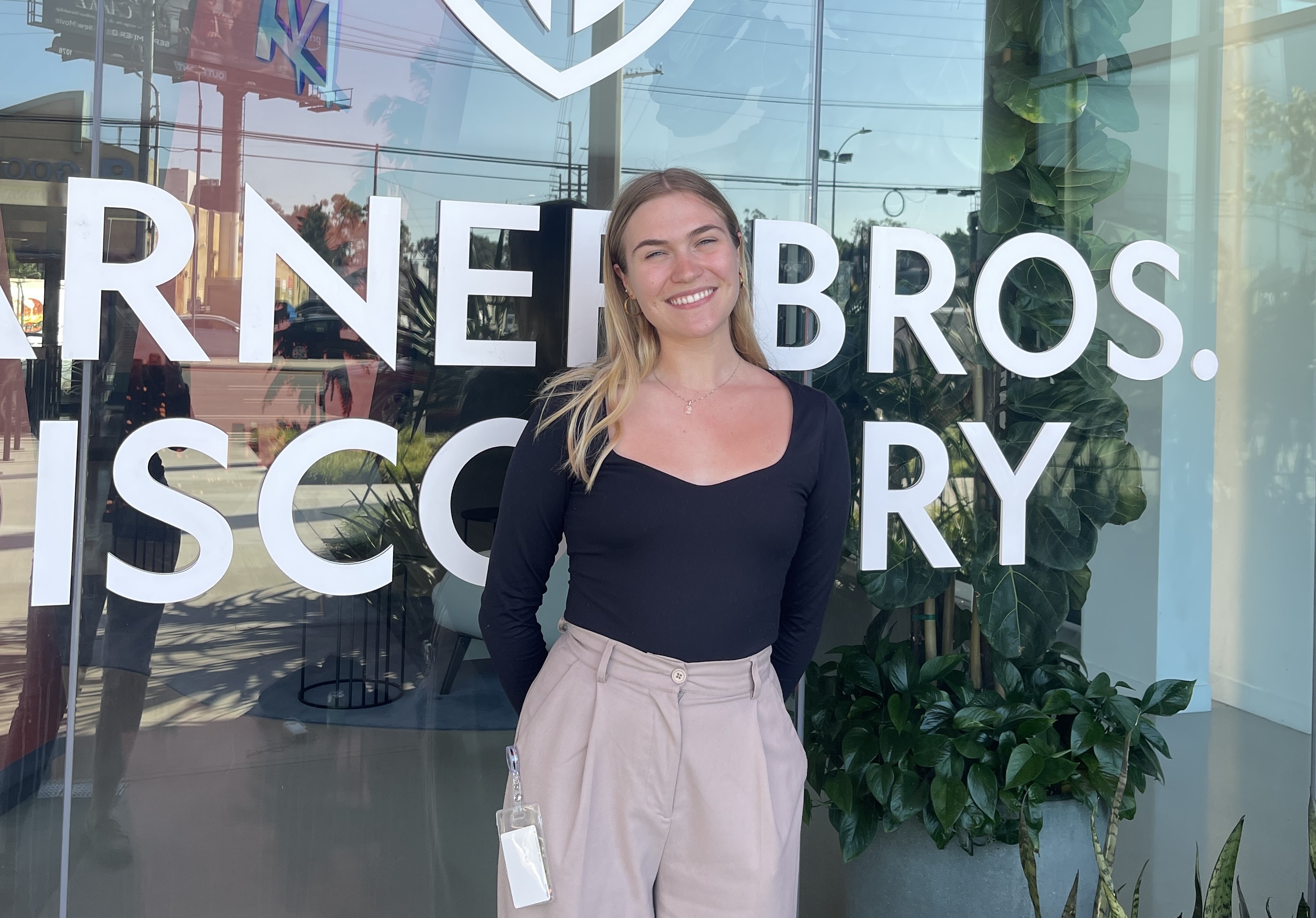 Headshot of student Riley Burke. She is pictured standing in front of a glass wall on the Warner Bros. Lot. She is smiling, wearing beige trousers, and a black long sleeve top.
