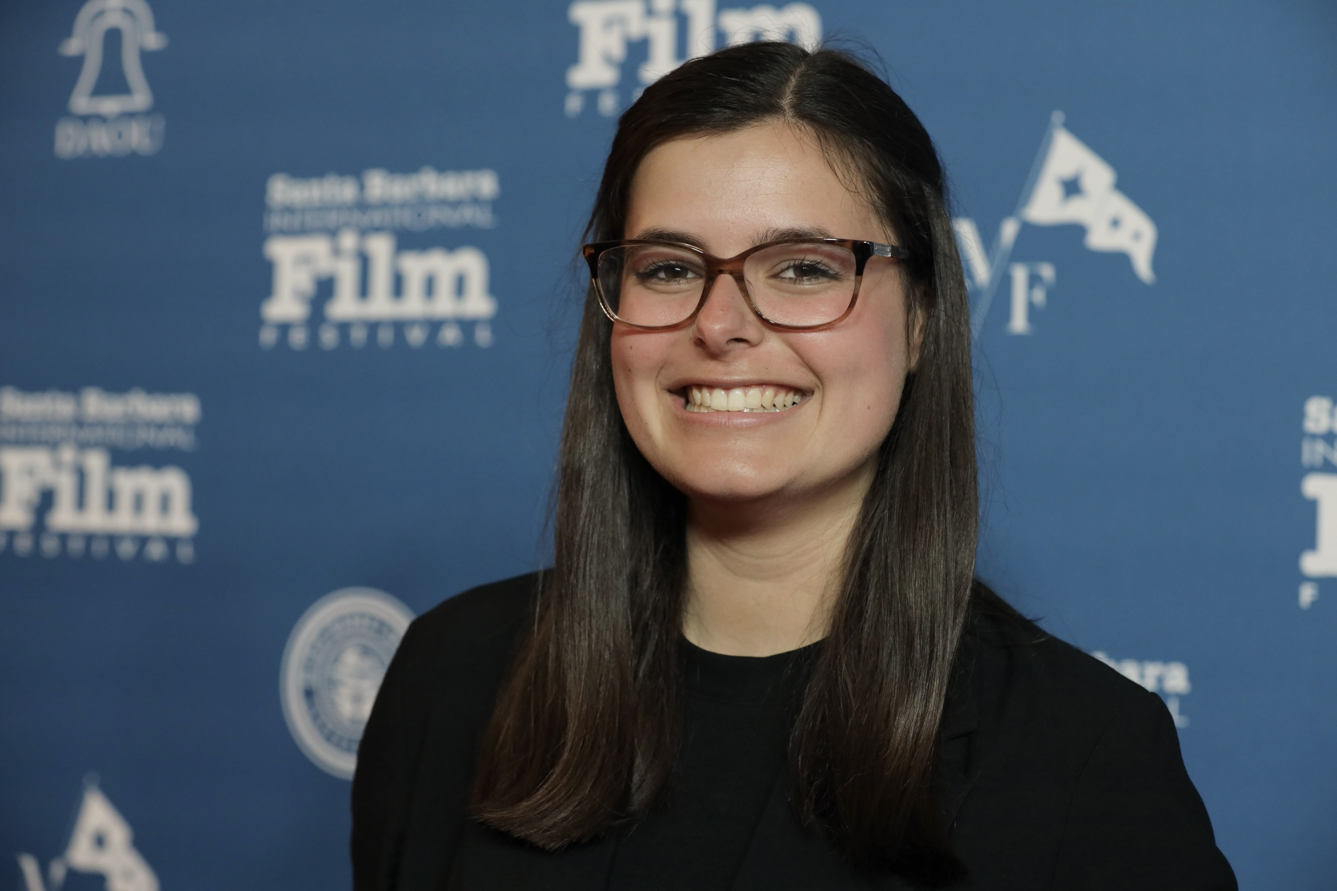 Red carpet headshot of Elle Viefhaus aon the closing Night of "Chosen Family" at the 39th Annual Santa Barbara International Film Festival. She is pictured swearing glasses, a black long sleeve shirt; she is smiling.