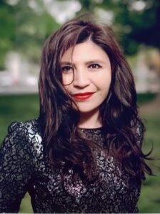 Headshot of activist Rayhan Asat. She is pictured smiling in front of a blurry park background, and is wearing a black and silver patterned top.