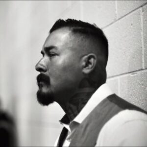 jerry-ramirez-headshot A headshot of Jerry Ramirez, documentary subject of the film American Homeboy. He is pictured leaning against a wall. Only his side profile can be seen. He dons a thick mustache and is wearing a white shirt and a dual-tone vest.