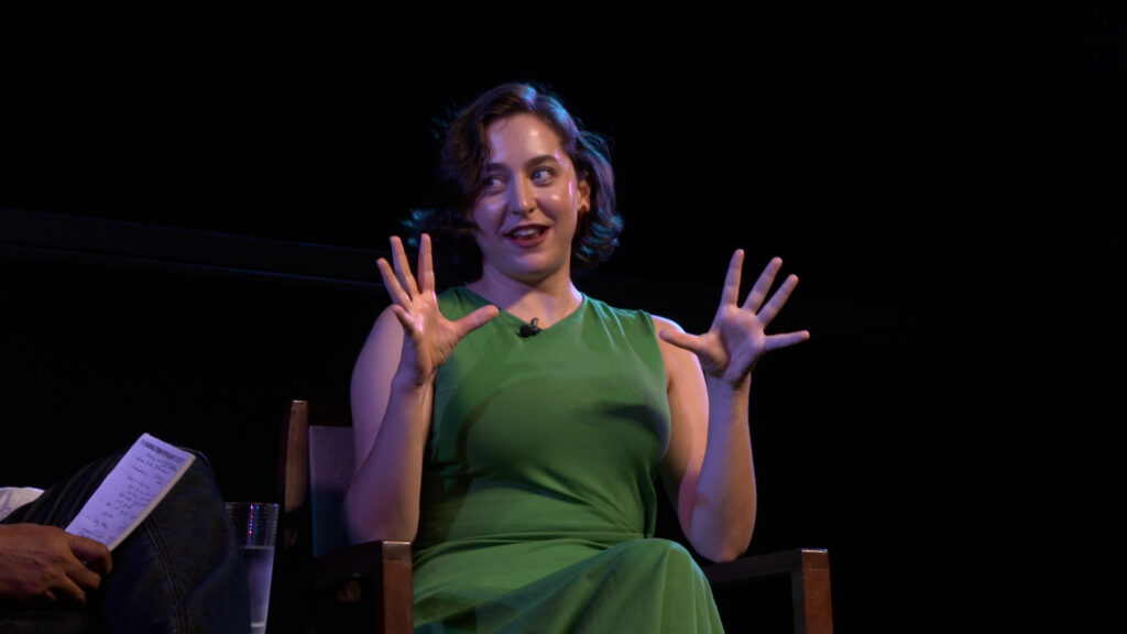 Stage photo of Sarah Friedland at the Pollock theater. She is pictured wearing a green dress. She is smiling and gesturing with both hands, as she looks off towards the moderator on her right off screen.