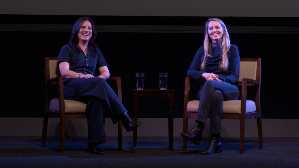 Stage photo of moderator Kelsey Moore and guest Charlotte Barker sat on stage at the Pollock Theater. Kelsey (left) appears smiling into the audience. Charlotte (right) sits smiling out into the audience.