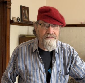 Filmmaker Gordon Quinn stands against a neutral background. He wears glasses, a beret, and a striped shirt.