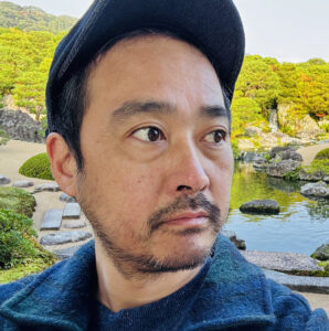 Film and Media Studies faculty member Naoki Yamamoto poses agains a background of trees and a pond. He is looking to the right, and wearing a blue jacket and cap.