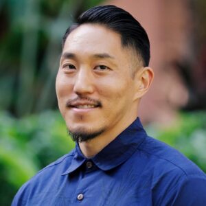 Tadashi Nakamura headshot Filmmaker Tadashi Nakamura stands against a leafy background. He is wearing a collared shirt and smiling at the camera.